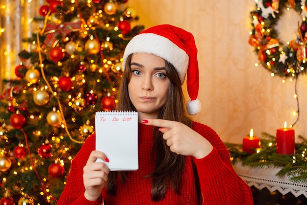 Concerned young girl in Santa hat and red sweater holding notebook and pointing on to do list phrase A lot of things to do before Christmas holidays