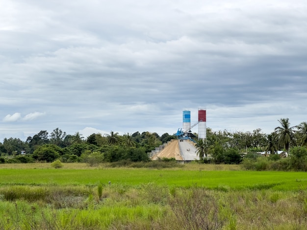 The concrete batching plant with the small crane is working in the construction site near the paddy field in the countryside area, front view with the copy space.