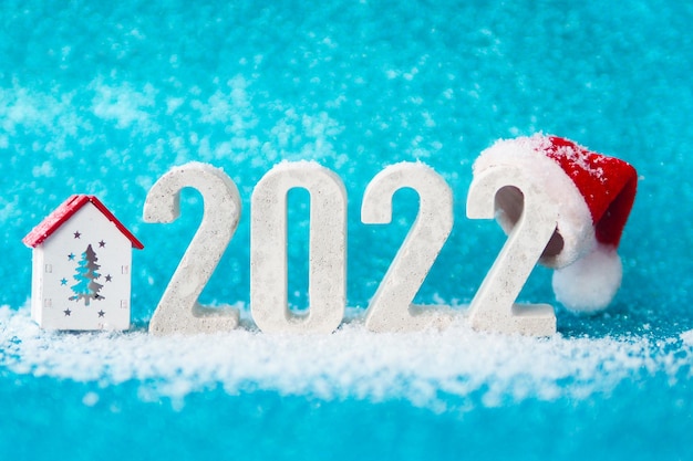 Concrete numbers 2022 with red Santa hat on blue glittering background and falling snow. copy space.