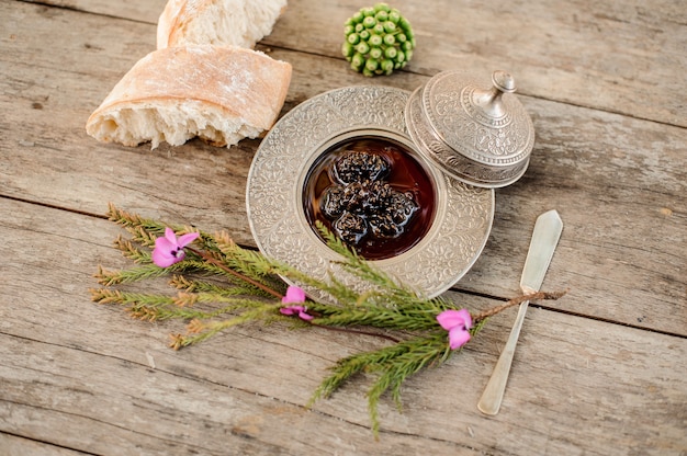 Cones jam in the silver plate with a cap, tree branch and bread near it on the wooden table