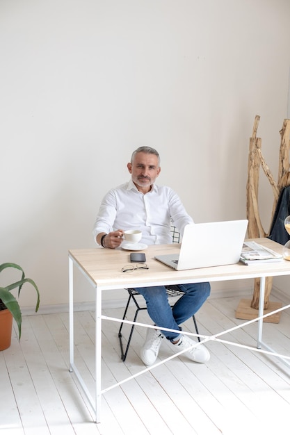 Confidence. Gray-haired business man with coffee sitting at table with laptop looking confidently at camera in daylight room