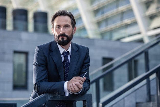 Confidence in mind. Waist up of a serious bearded businessman leaning on the handrail while looking at you