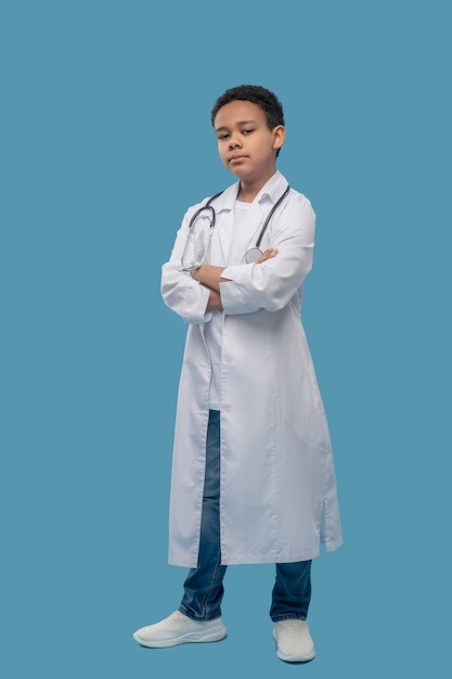 Confidence. Serious black boy of primary school age standing in white medical coat with folded hands on chest against blue background