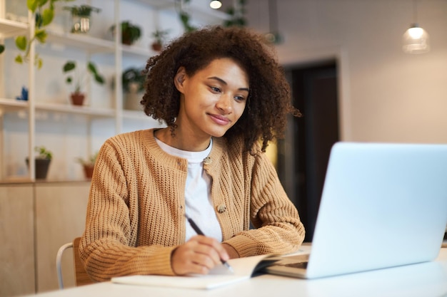 Confident African American young woman smart student making notes while watching webinar on laptop