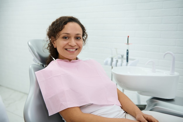 Confident African woman sitting in dentist's chair, ready to receive dental treatment at medical check-up, smiles toothy smile looking at camera. Dentistry, early prevention of teeth diseases concept