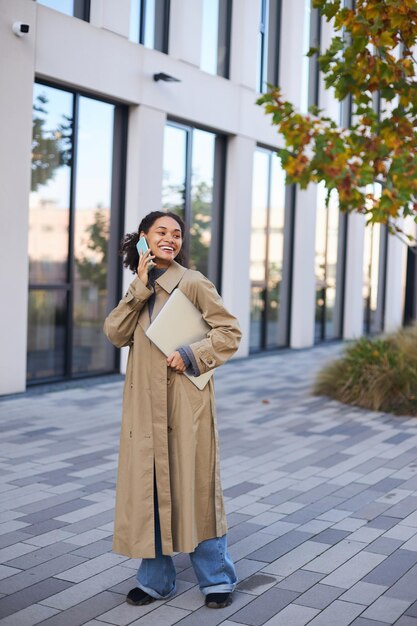 Confident African woman talking on mobile phone outside the office building with a laptop in hands