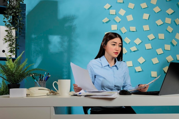 Confident Asian business woman doing paperwork while sitting at office desk. Focused entrepreneur using laptop with papers in hand preparing a report analyzing work results.