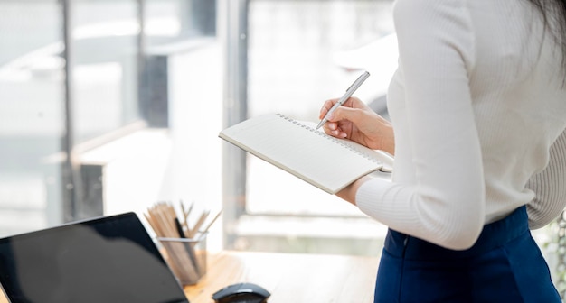 Confident Asian businesswoman standing and taking notes in financial book Income tax and working on laptop at office