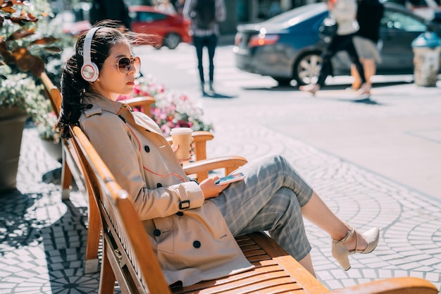 confident asian chinese woman in sunglasses listening to music from smartphone with headphones in city street. young girl with headset enjoy sunshine sitting on wood bench holding mobile phone.