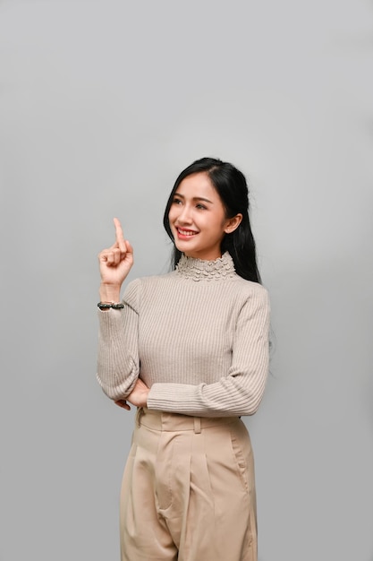 Confident Asian female smiling pointing up her finger standing against the grey wall