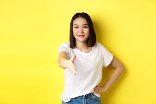 Confident asian woman in white t-shirt, stretch out hand for handshake and greeting gesture, saying hello, introduce herself, standing over yellow.