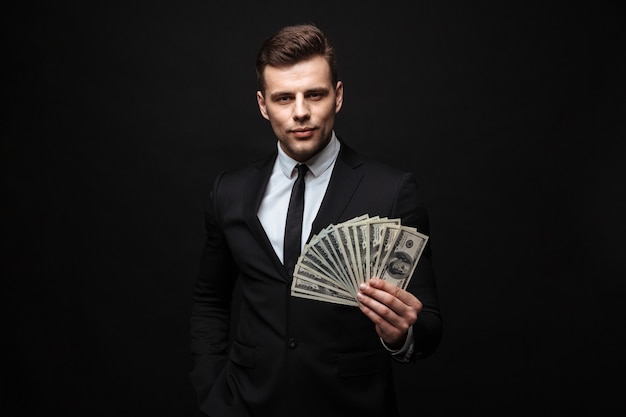 Confident attractive young businessman wearing suit standing isolated over black wall, showing money banknotes