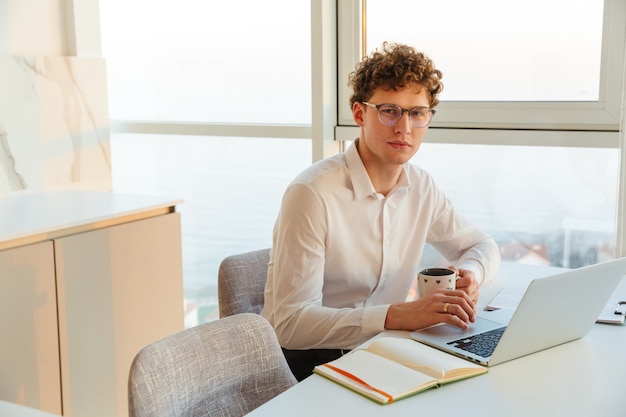 Confident attractive young businessman wearing white shirt working on laptop computer while sitting at the table at home, drinking coffee from a mug