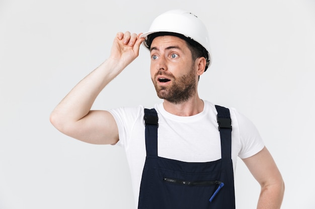 Confident bearded builder man wearing overalls and hardhat standing isolated over white wall, looking away