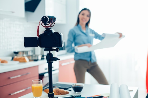 Confident blogger standing in the kitchen with roll of architectural paper and recording a video with a help of a camera on her table