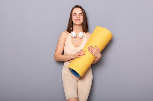 Confident brown haired woman holding yoga mat isolated over gray background looking at camera training her body prefers healthy active lifestyle