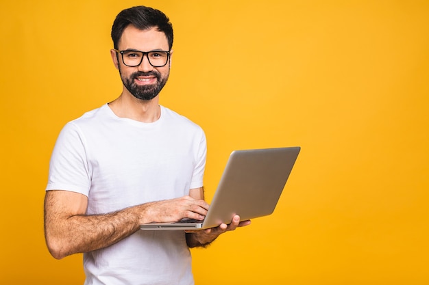 Confident business expert. Confident young handsome bearded man in casual holding laptop and smiling while standing over isolated yellow background.