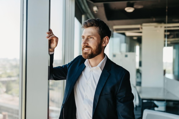 Confident business man in suit standing in modern coworking office room leaning on window and smiling