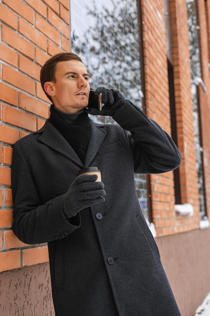 Confident businessman in coat with paper cup of takeaway coffee speaking on cellphone in city