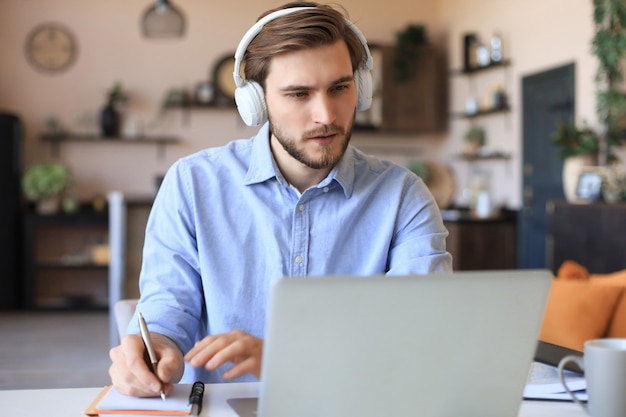 Confident businessman in earphones is writing notes or financial report while sitting at desk with laptop at home.