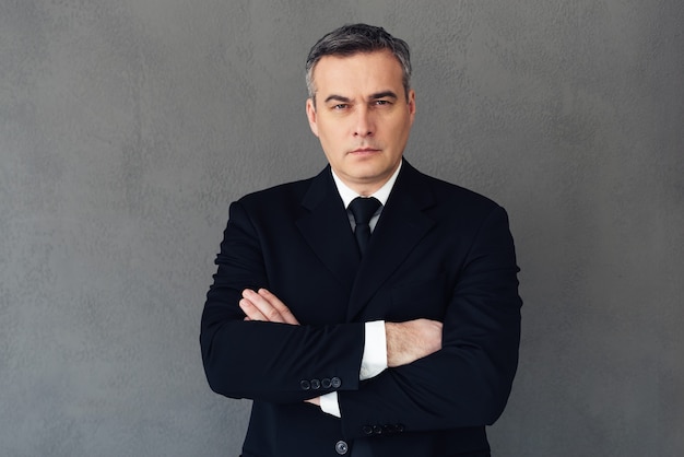 Confident businessman. Mature businessman keeping arms crossed and looking at camera while standing against grey background