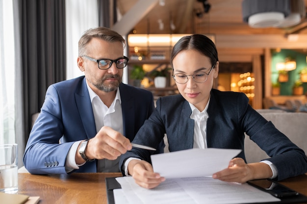 Confident businessman pointing at contract while his female colleague reading its terms and points at meeting in restaurant