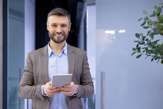 Confident businessman in smart casual attire smiling holding a tablet in modern office setting