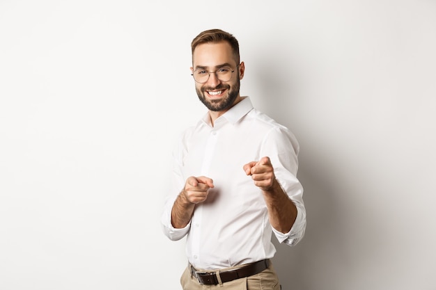 Confident businessman smiling, pointing fingers at you, congrats or praise gesture, standing over white background.