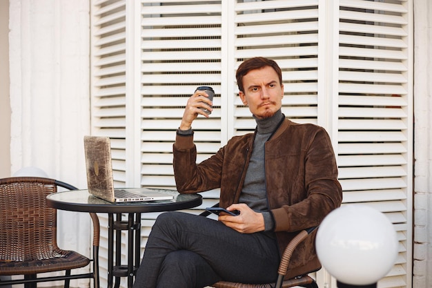 Confident businessman with cup of coffee using mobile phone and having break in street cafe in city