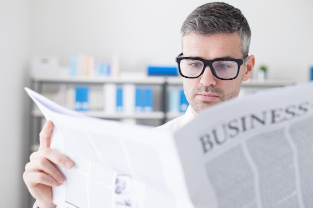 Confident businessman with glasses reading a financial newspaper in his office