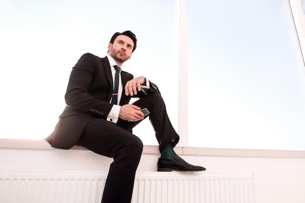 Confident businessman with a smartphone sitting in the office hallway