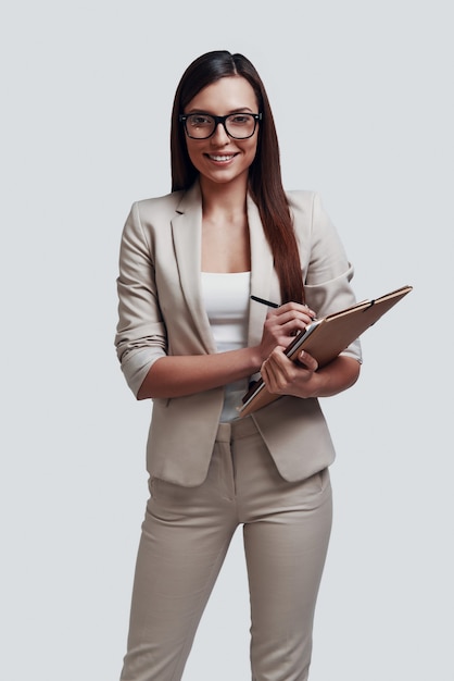 Confident businesswoman. Attractive young woman looking at camera and smiling while standing against grey background