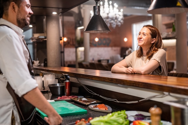 Confident chef standing at counter of open kitchen and talking to restaurant guest