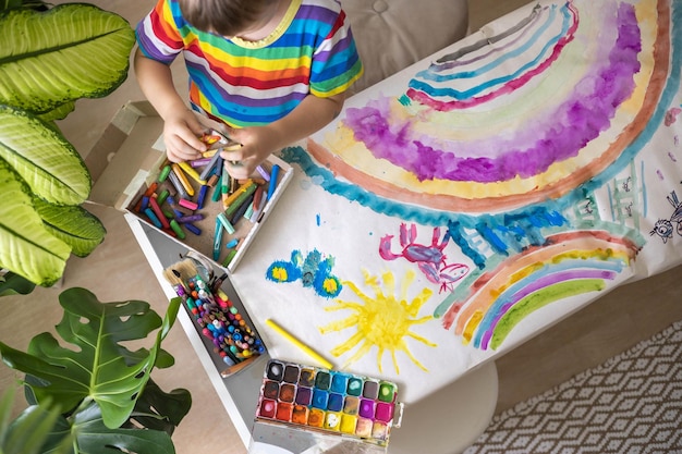 Photo confident cute male kid art drawing multicolored rainbow arch use paints and brush on table at kindergarten home interior focused baby boy enjoying artwork early development creative activity