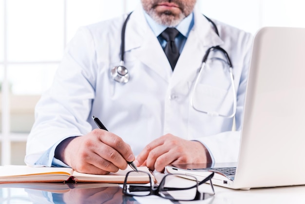 Confident doctor at work. Close-up of mature doctor writing something in note pad while sitting at his working place