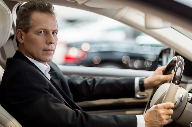 Confident drive. Side view of confident senior man in formalwear sitting in car and looking at camera