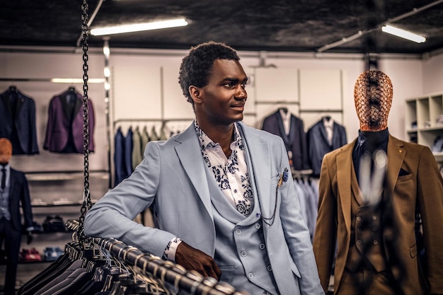 A confident elegantly dressed African-American man leaning on a mannequin in a classic menswear store.