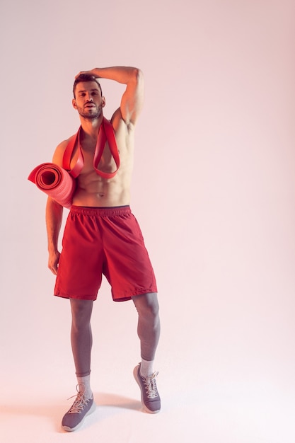 Confident european sportsman posing with resistance band and fitness mat. Young handsome bearded man with naked sportive torso. Isolated on beige background. Studio shoot. Copy space