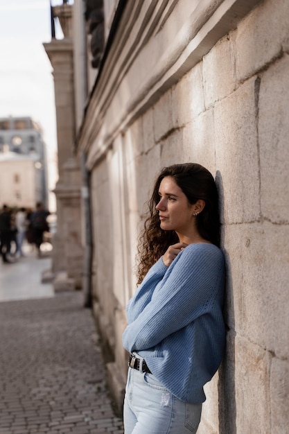 Confident girl model weared in blue pullover is posing outdoor in city Portrait of attractive business woman with curly hair is posing on grey wall background