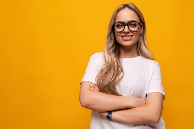 Confident girl student in glasses and a white tshirt crossed her arms on a yellow background with