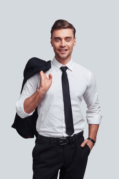 Confident and handsome. Good looking young man carrying his jacket on shoulders and smiling while standing against grey background