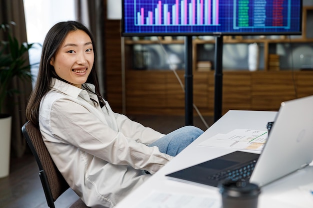 Confident happy smiling diverse asian woman startup team leader He sits at his workplace with a laptop coffee on the background of a TV with charts and an analytical summary of marketing strategy