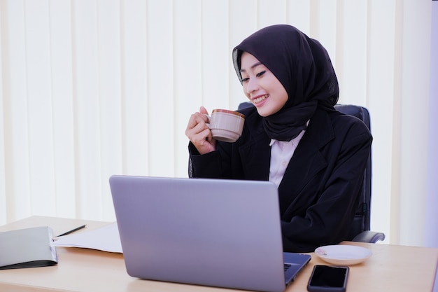 Confident happy young business woman sitting at office desk, holding a cup of coffee