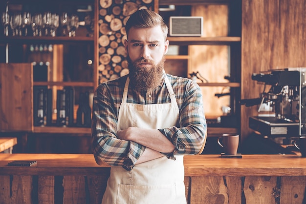 Confident in his new business. Young bearded man in apron looking at camera and keeping arms crossed while standing at bar counter