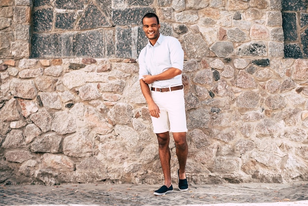 Confident in his perfect style. Full length of cheerful young African man in smart casual clothes adjusting his shirt sleeve and looking at camera while standing against the stoned wall outdoors