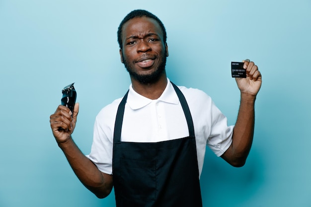 Confident holding card with hair clippers young african american barber in uniform isolated on blue background