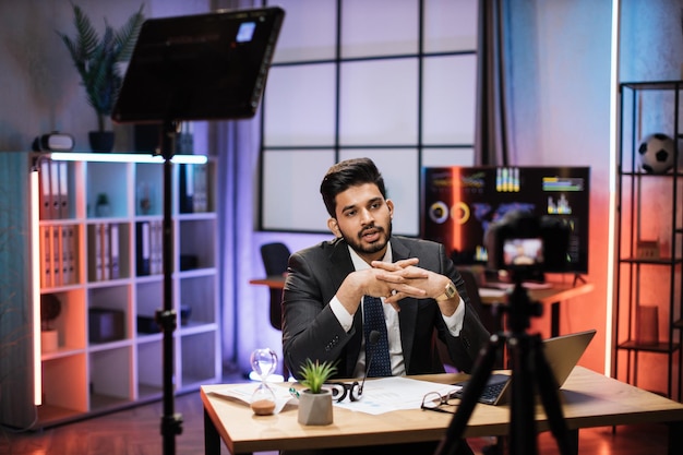 Confident indian bearded businessman in suit sitting in front of camera in evening office