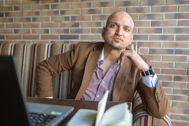 Confident Indian man sitting at cafe with notebook and laptop with seriously looking at camera