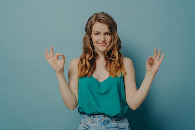 Confident joyful young woman in casual clothes gesturing ok sign with both hands in studio