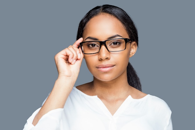 Confident look. Attractive young African woman adjusting her eyeglasses and looking at camera while standing against grey background